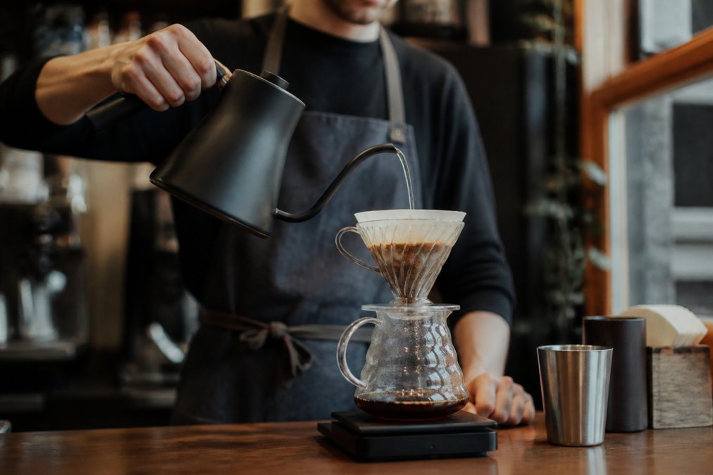 Close-up of a barista pouring water from a gooseneck kettle in a slow, steady spiral over coffee grounds in a pour-over brewer, with a glass carafe below, warm morning light from a window