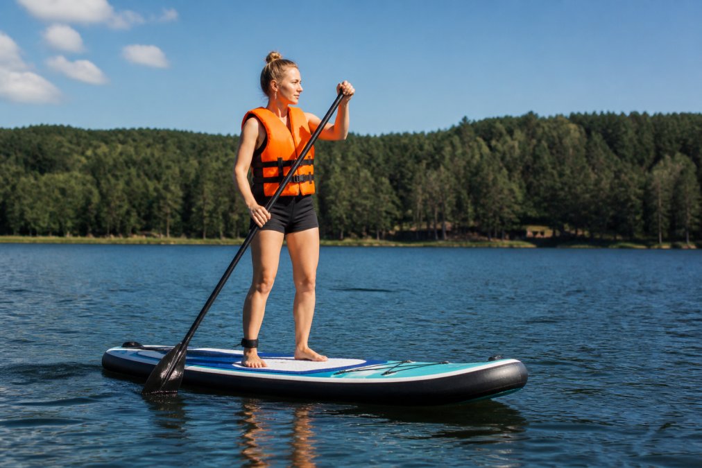 Beginner paddleboarder standing upright on an inflatable stand-up paddleboard on calm lake water, wearing a bright orange personal flotation device and holding a paddle, with forested shoreline visible in the distance