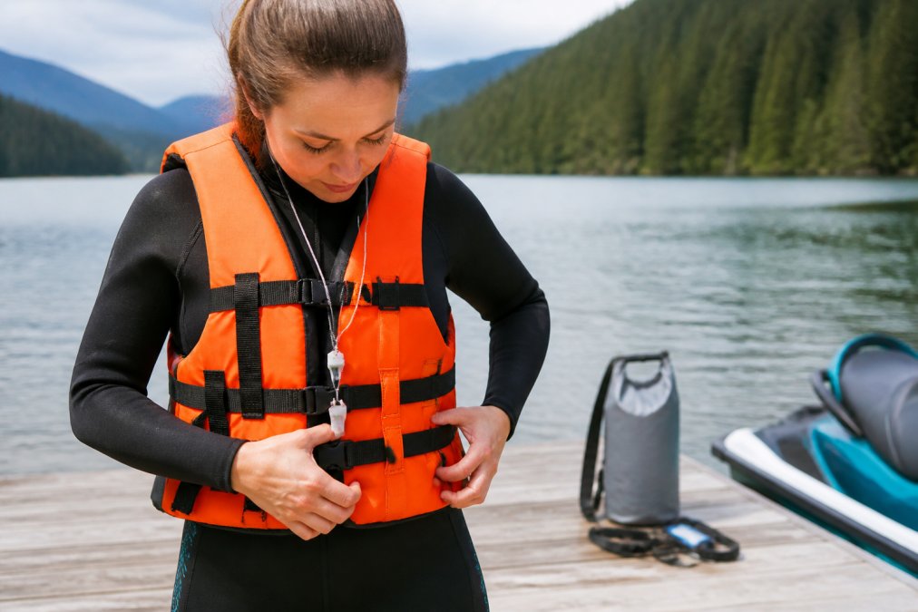 Person in water sports gear carefully adjusting and checking their orange life vest with hands, wearing a whistle on a lanyard around their neck, with safety equipment like a dry bag and leash visible on a dock in the background