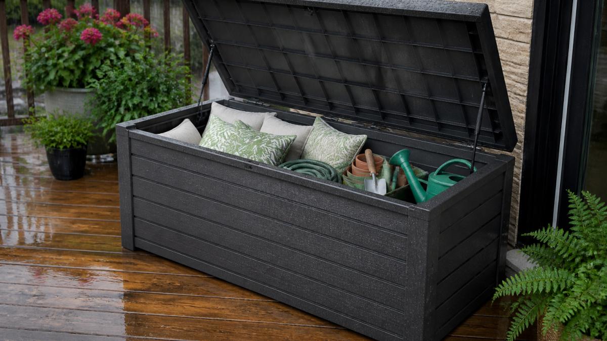Open waterproof deck storage box on a patio during light rain, showing interior organization with cushions and garden tools neatly stored, water beading on the sealed lid