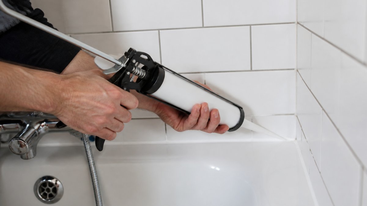Professional tradesperson's hands applying clear silicone sealant to a bathroom tile joint using a caulking gun, with white subway tiles and grey grout visible in natural bathroom lighting