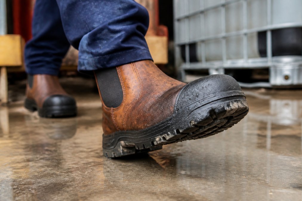 Close-up of tradesperson's foot wearing waterproof slip-on work boot on wet concrete surface, showing slip-resistant tread pattern and steel toe cap in action