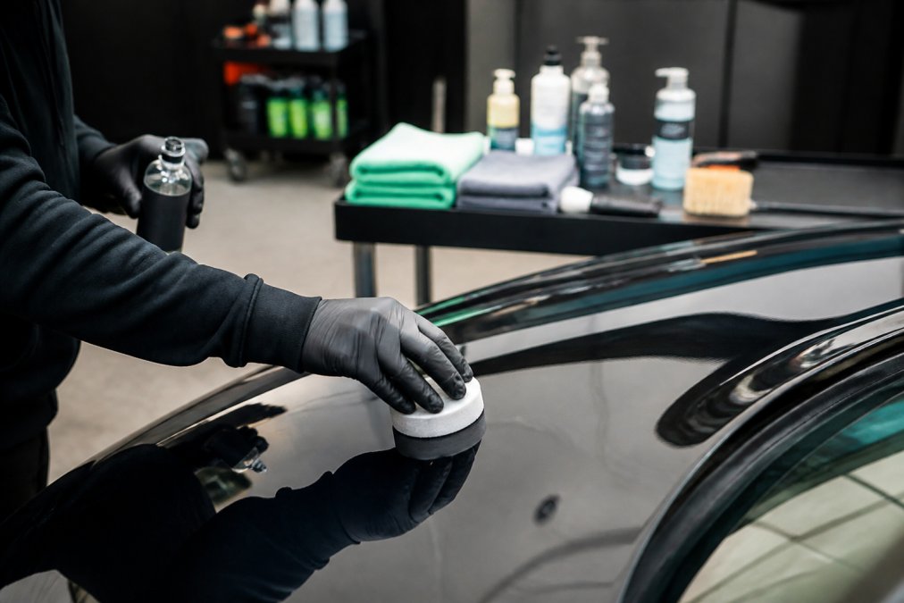 Handen in werkhandschoenen die synthetische sealant aanbrengen op autolak met applicatorpad, microvezeldoeken en detailingproducten zichtbaar op werkbank in professionele garage