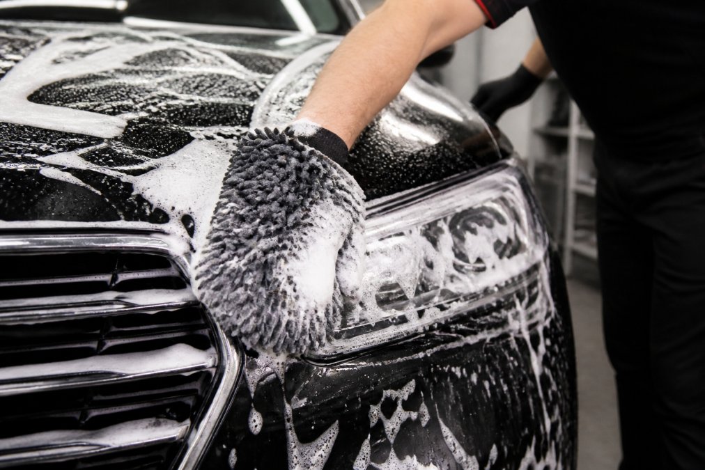 Close-up van iemand die een auto wast met washandschoenen en auto shampoo, zichtbare schuimlaag op de voorkant van een voertuig