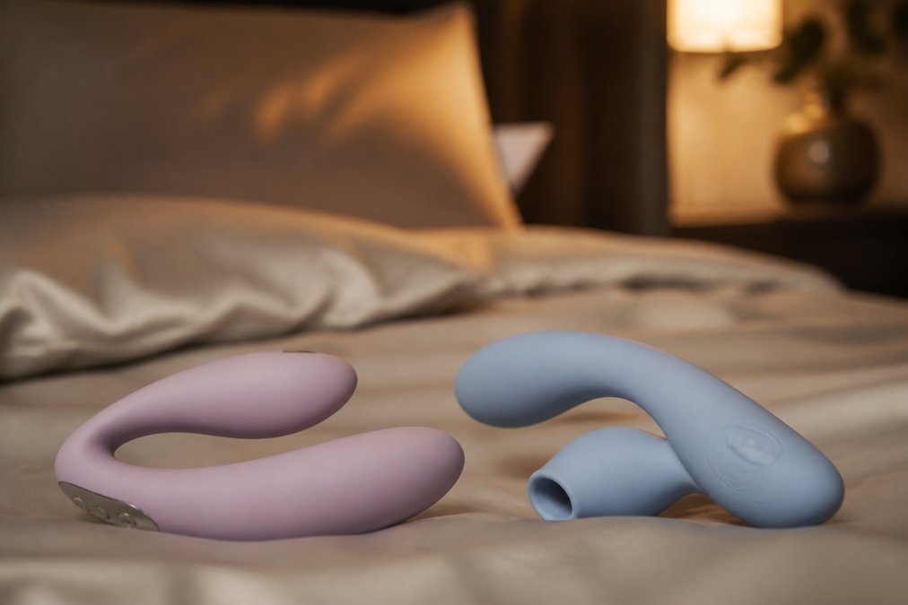 Twee moderne, ergonomische koppelvibrators in zachte pasteltinten liggen op satijnen beddengoed, warme gedempte verlichting, luxe slaapkamer setting