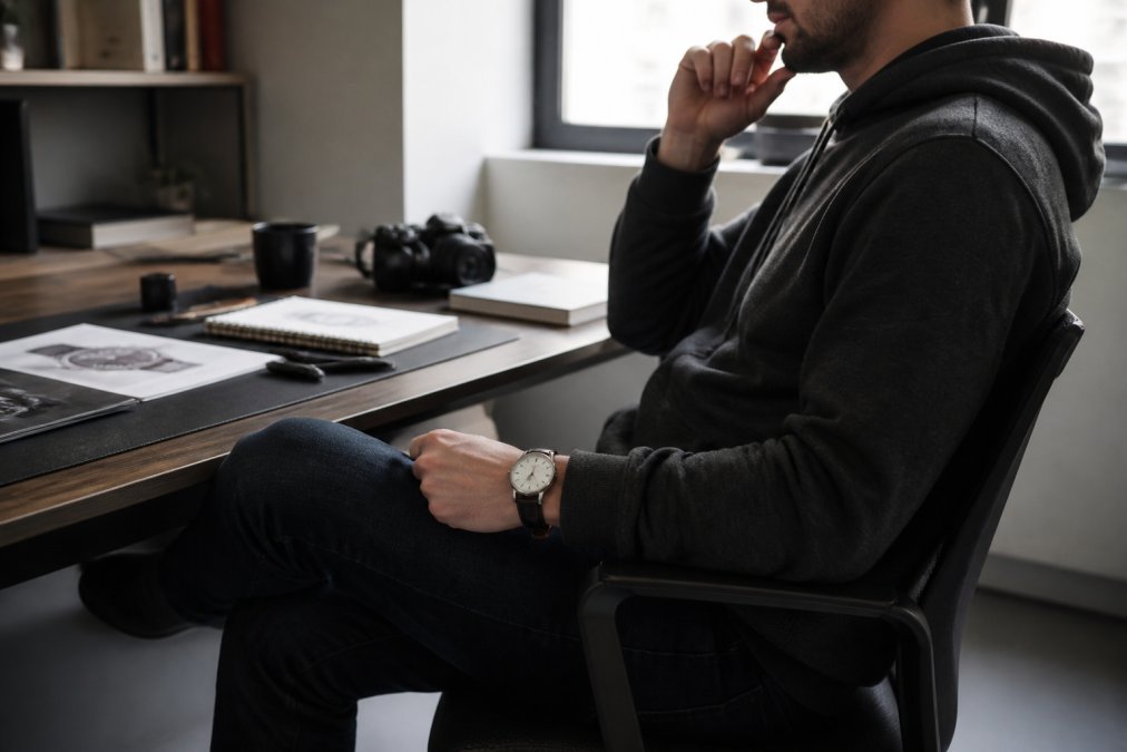 A person in a charcoal hoodie and dark denim jeans sitting at a minimalist desk, wearing a slim dress watch with a leather strap, natural window light, shallow depth of field