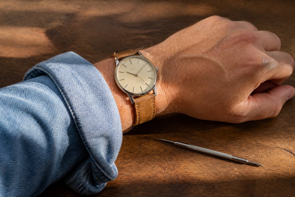 Close-up of a wrist wearing a minimalist dress watch with a tan leather strap, paired with a rolled-up denim shirt sleeve, a spring bar tool resting on a wooden table nearby in warm afternoon light