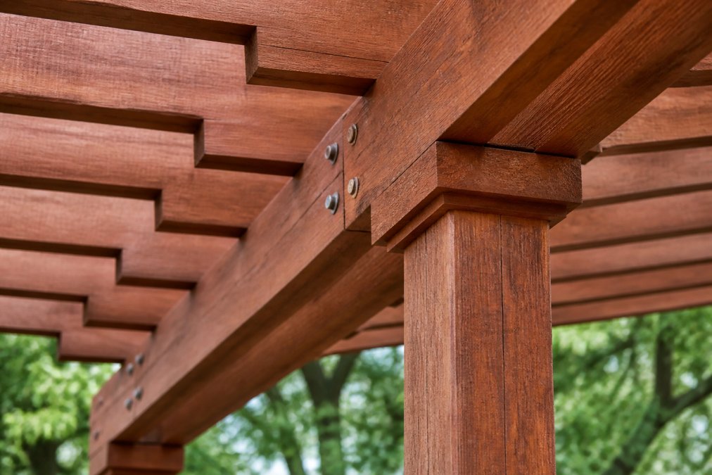 Close-up of natural cedar wood pergola showing warm reddish-brown grain texture and visible weathering patterns from outdoor exposure, with soft natural lighting highlighting the wood's depth