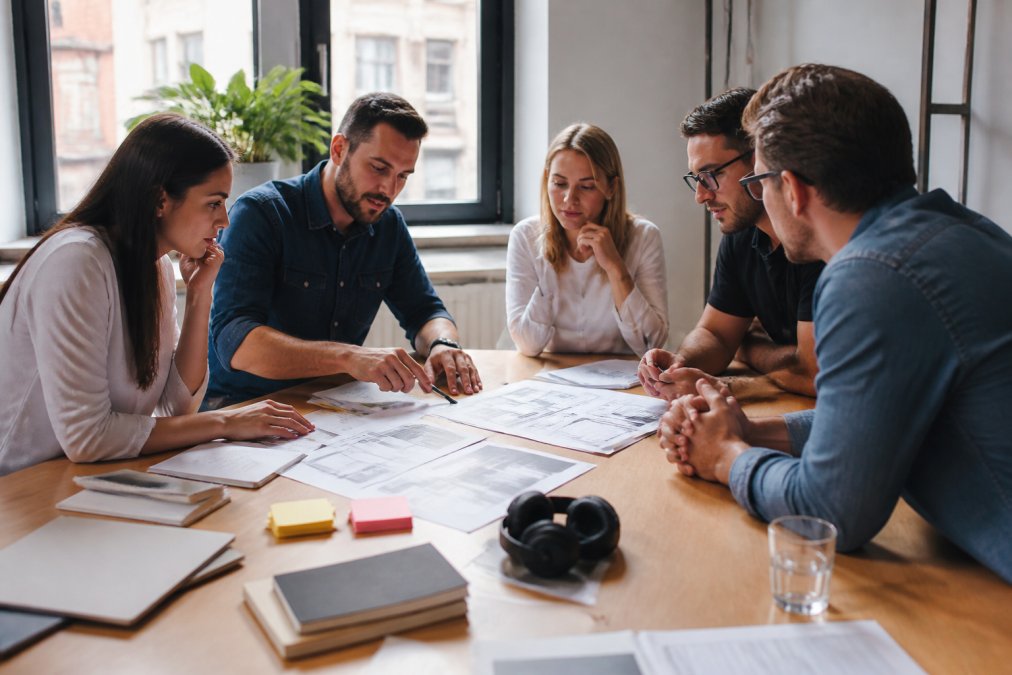 Web development team collaborating in modern office environment with developers and designers reviewing code and design mockups on multiple screens under bright natural lighting