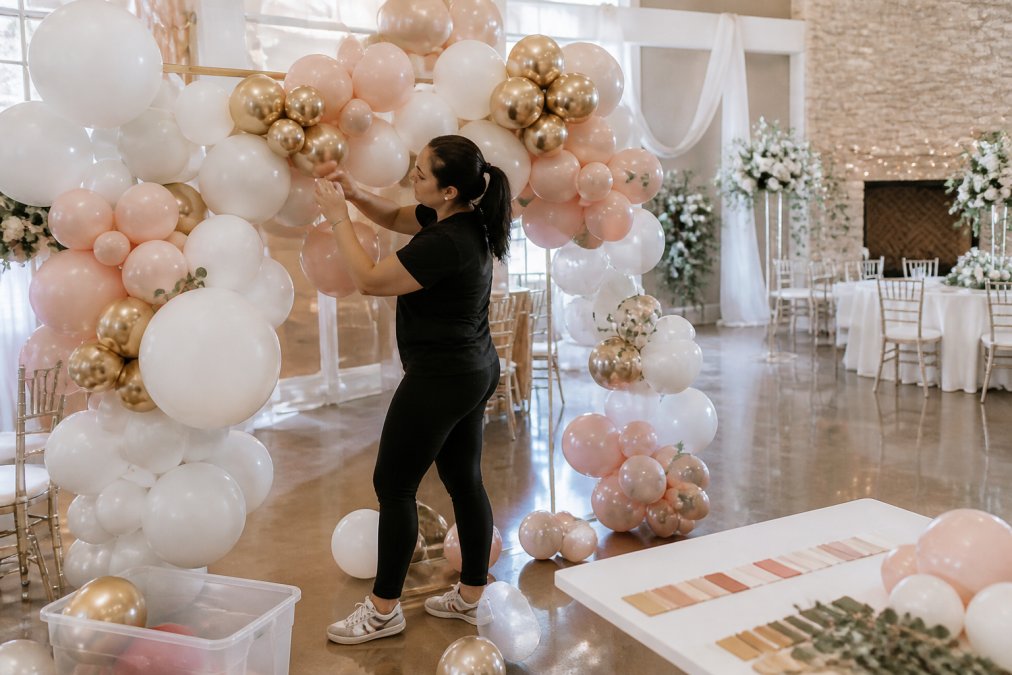 A professional balloon artist in a bright studio assembling a large organic balloon garland in white, blush, and gold tones on a frame, with a wedding venue backdrop visible in the background and a color palette board nearby