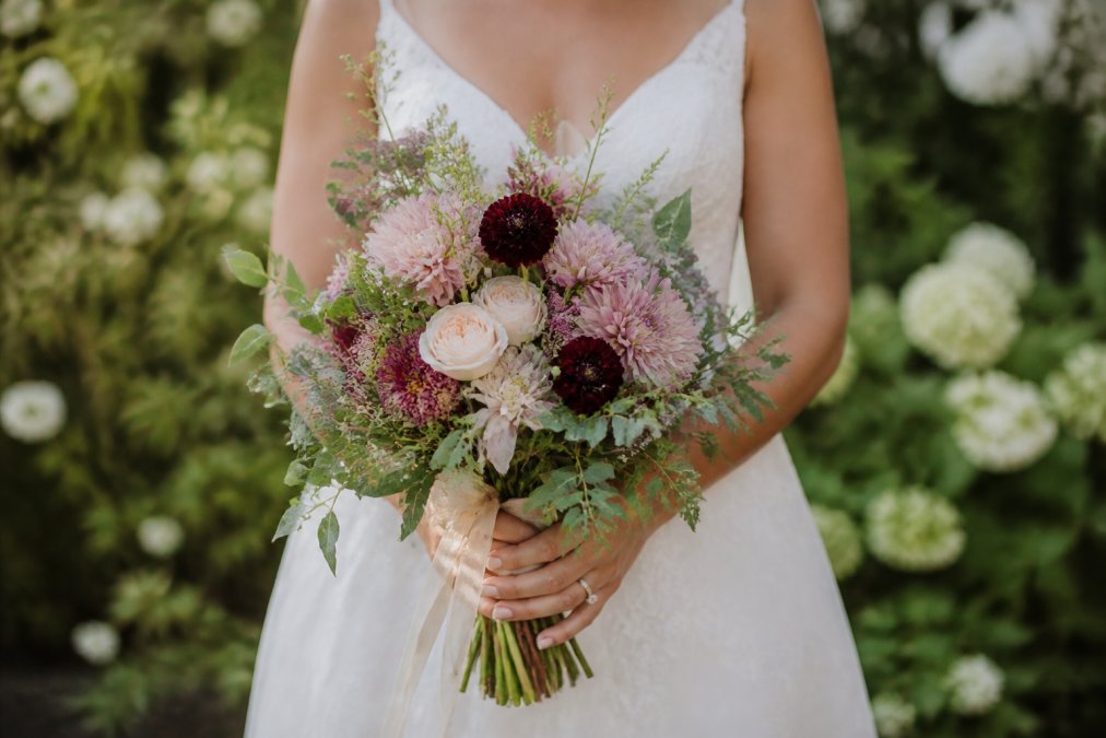 Bride holding a loose, texture-rich hand-tied wedding bouquet of fresh seasonal blooms in a garden setting