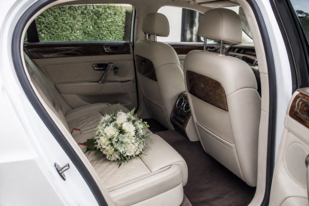 The interior of a luxury white Bentley Flying Spur showing cream leather seats, wood veneer detailing, and a bridal bouquet resting on the rear seat