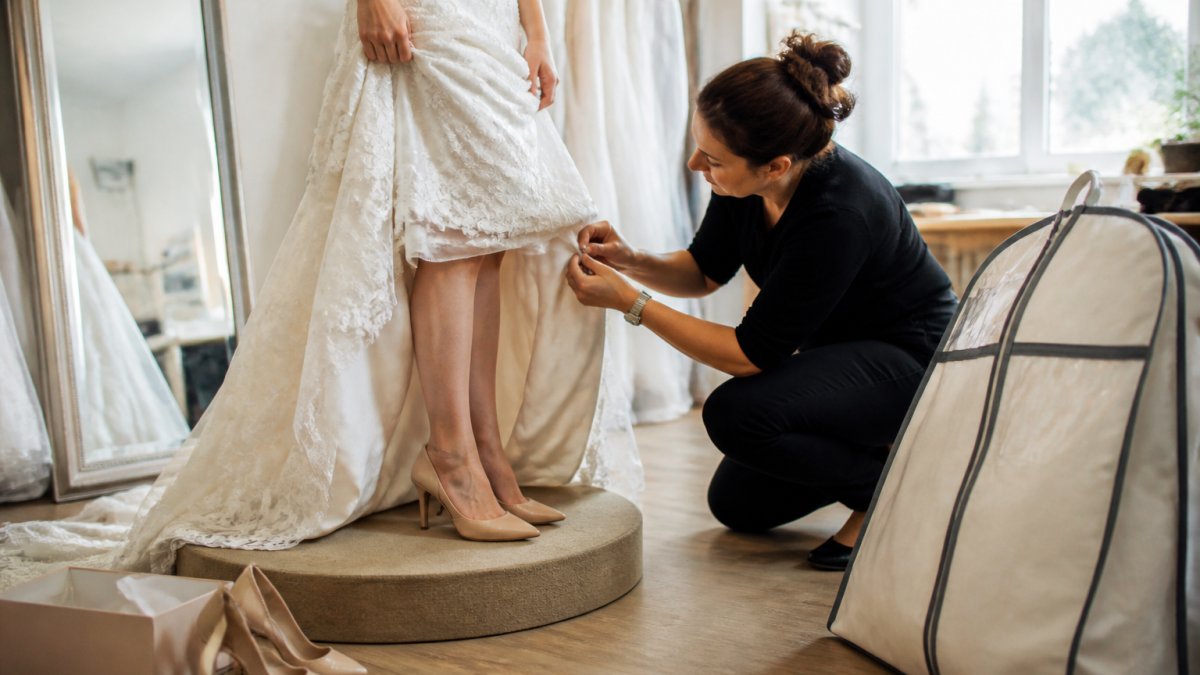 A bride standing on a fitting pedestal in a bridal salon, wearing her wedding shoes and undergarments while a seamstress pins the hem of her dress, with a garment bag and shoe box nearby