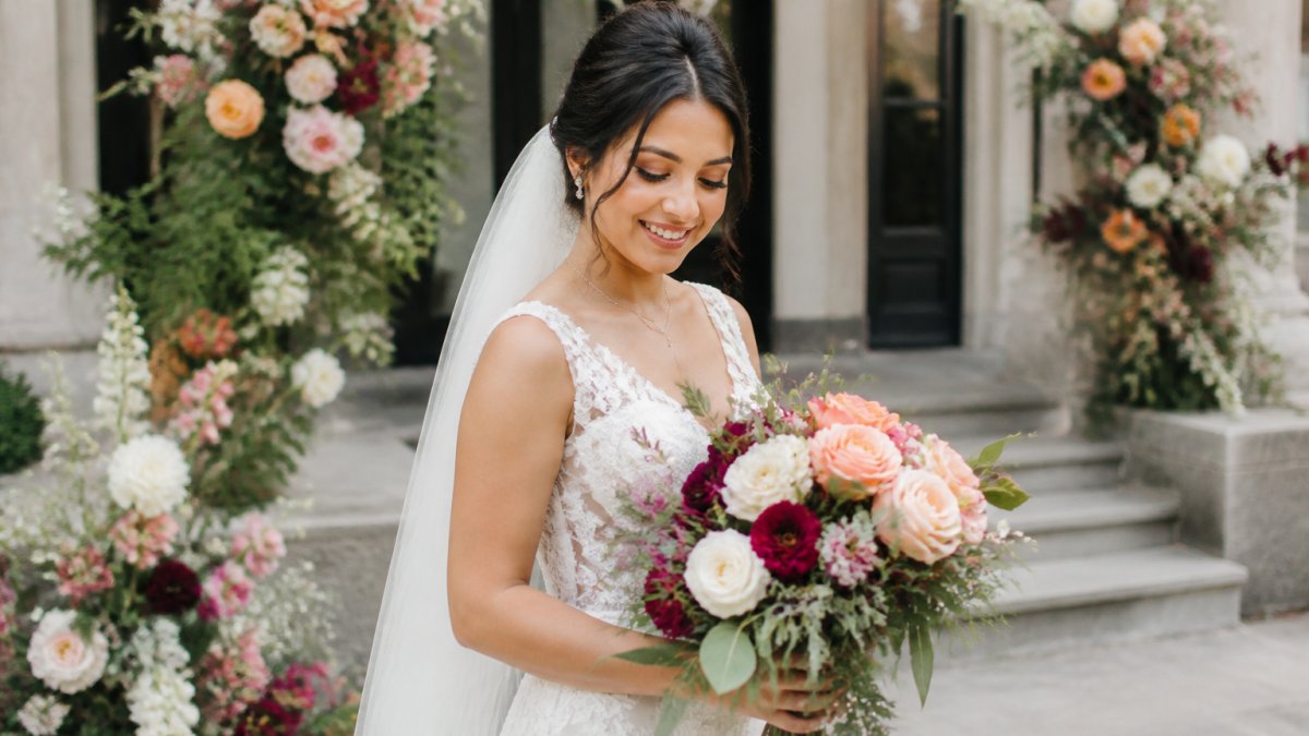 Bride holding a vibrant bridal bouquet with seasonal flowers standing in front of a wedding venue with floral arrangements visible in the background, natural outdoor lighting