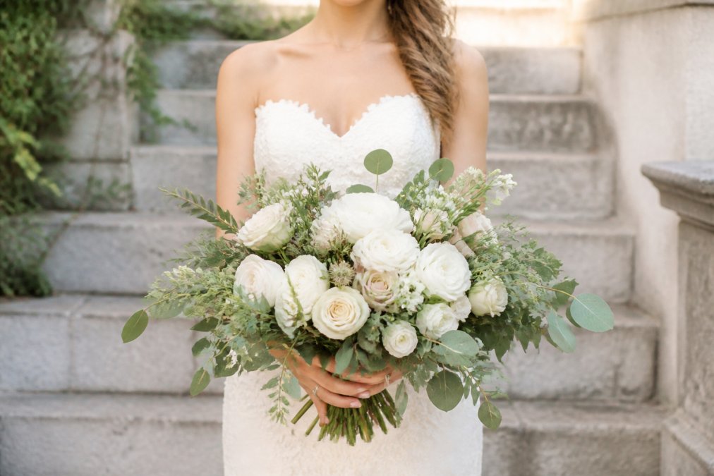 Bride holding a lush bridal bouquet with focal flowers and greenery, standing in natural light to show floral arrangement detail and quality