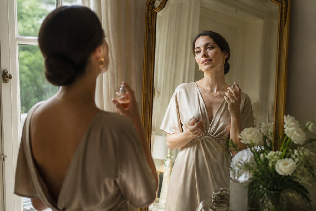 A well-dressed woman in her 30s standing in front of a mirror, holding a perfume bottle, with soft natural light coming through a window, creating a warm and reflective atmosphere