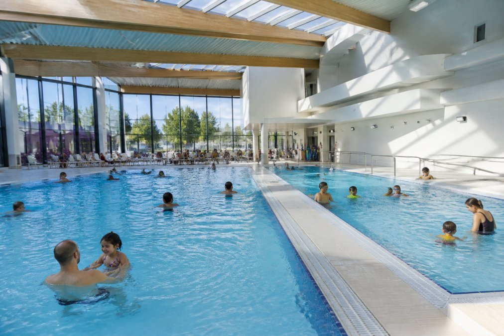 Adults and children swimming in a bright indoor pool with overhead skylights and modern architectural features, some in the main pool and others in a teaching pool, with poolside seating visible