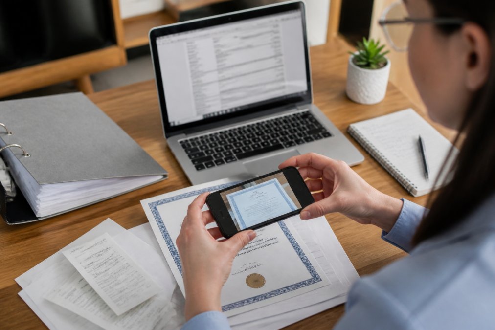 Nahaufnahme von Händen, die Rechnungen und Kurszertifikate mit einem Smartphone scannen, während auf dem Schreibtisch ein Laptop mit offener Steuererklärung steht und Notizen daneben liegen