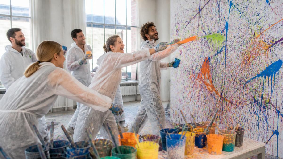 Eine Gruppe Erwachsener in Schutzkleidung wirft lachend Farbe auf eine große Leinwand in einem hellen Atelier, im Hintergrund liegen Pinsel und Farbbecher bereit