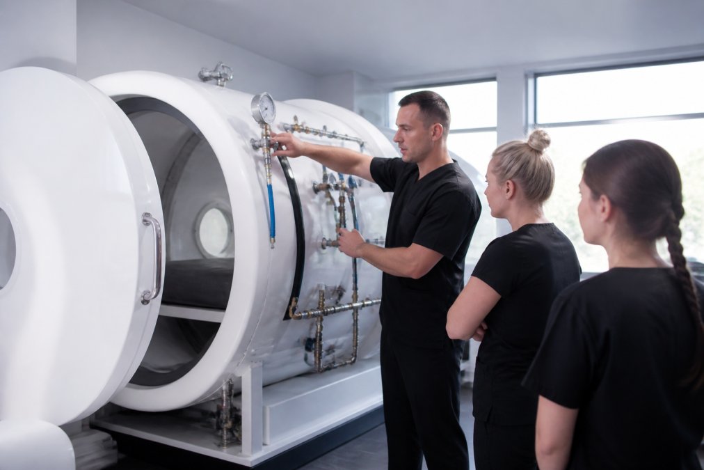Professional wellness clinic staff member conducting equipment training and demonstrating proper maintenance procedures on hyperbaric chamber in modern clinical setting with bright overhead lighting