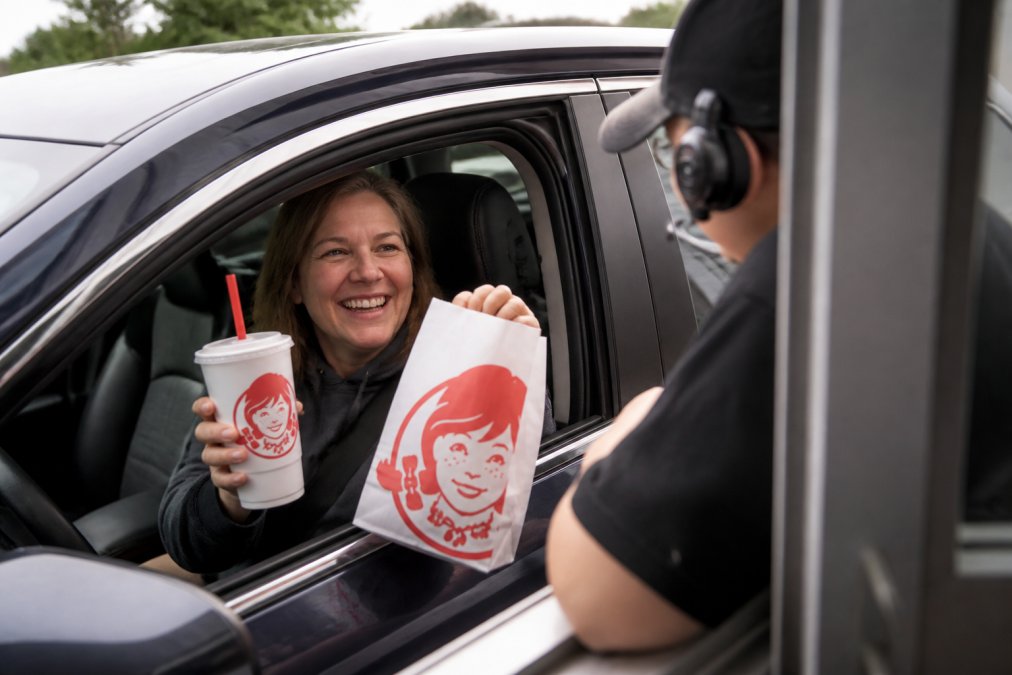 Satisfied customer in car holding Wendy's order bag and drink, smiling at drive-thru window during daytime