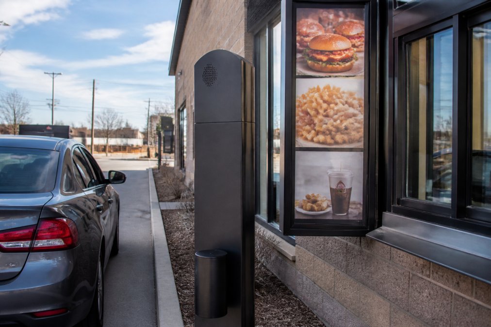 Modern Wendy's drive-thru window with digital menu boards and AI ordering screen interface visible, customer vehicle approaching the ordering speaker, bright daylight illuminating the service area