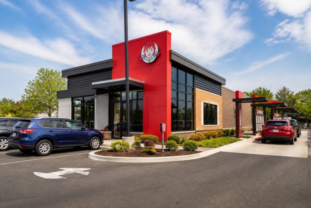 Exterior view of a Wendy's restaurant location with red signage, drive-thru lane visible, parking area with customer vehicles, modern storefront design