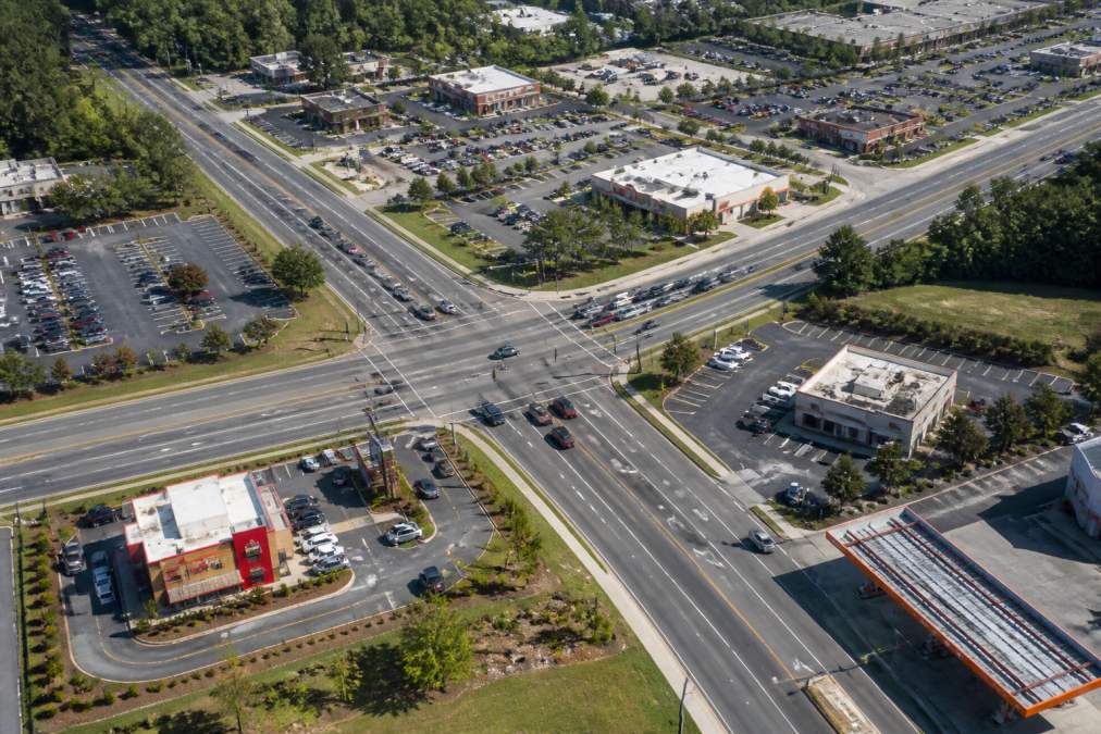 Aerial view of a busy commercial intersection with multiple retail establishments, parking lots, and traffic patterns visible, representing market density and site selection analysis