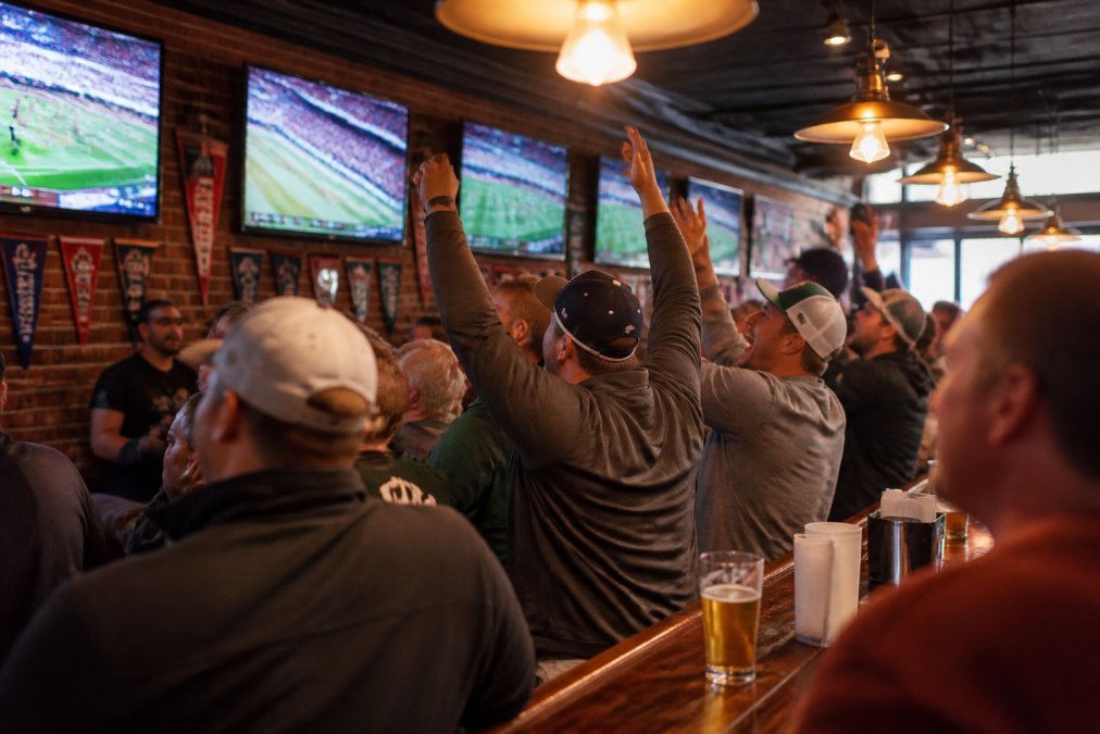 A lively sports bar interior with a large crowd watching a game on multiple high-definition televisions, bar counter in the foreground, warm overhead lighting and team pennants on the wall