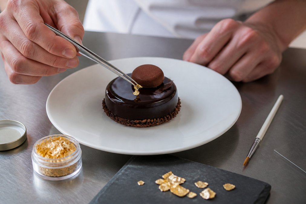 Hands using tweezers to place delicate authentic gold flakes onto a decadent chocolate dessert on a white plate.