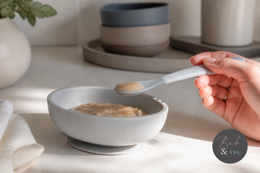 Close-up of a parent's hand holding a soft silicone baby spoon next to a bowl of puree on a clean kitchen counter, natural morning light