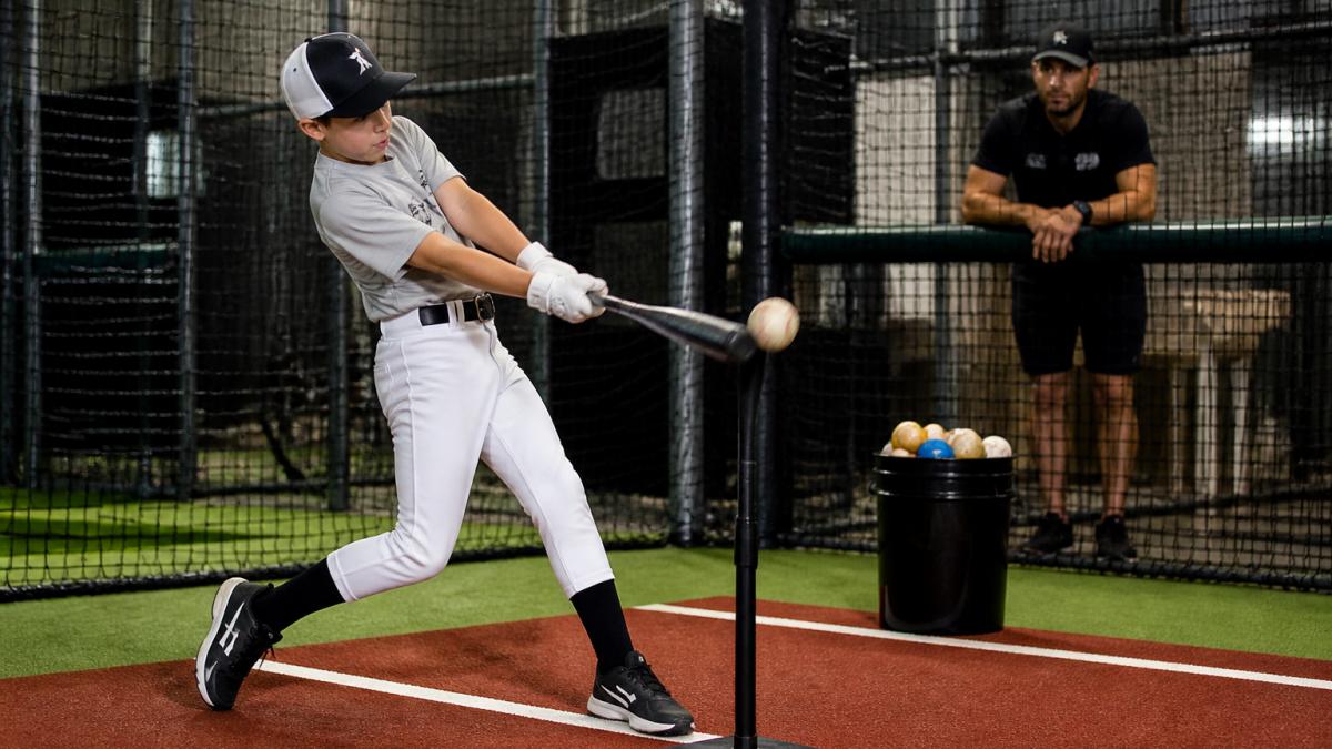 Youth baseball player mid-swing in a batting cage, hitting a practice baseball off a tee, with a bucket of assorted practice balls nearby and a coach watching from behind the net