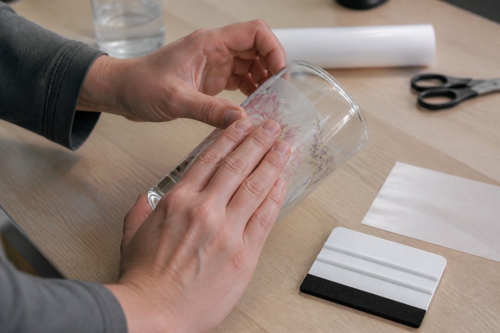 Hands using a squeegee to apply UV DTF decals to a glass tumbler on a clean workbench