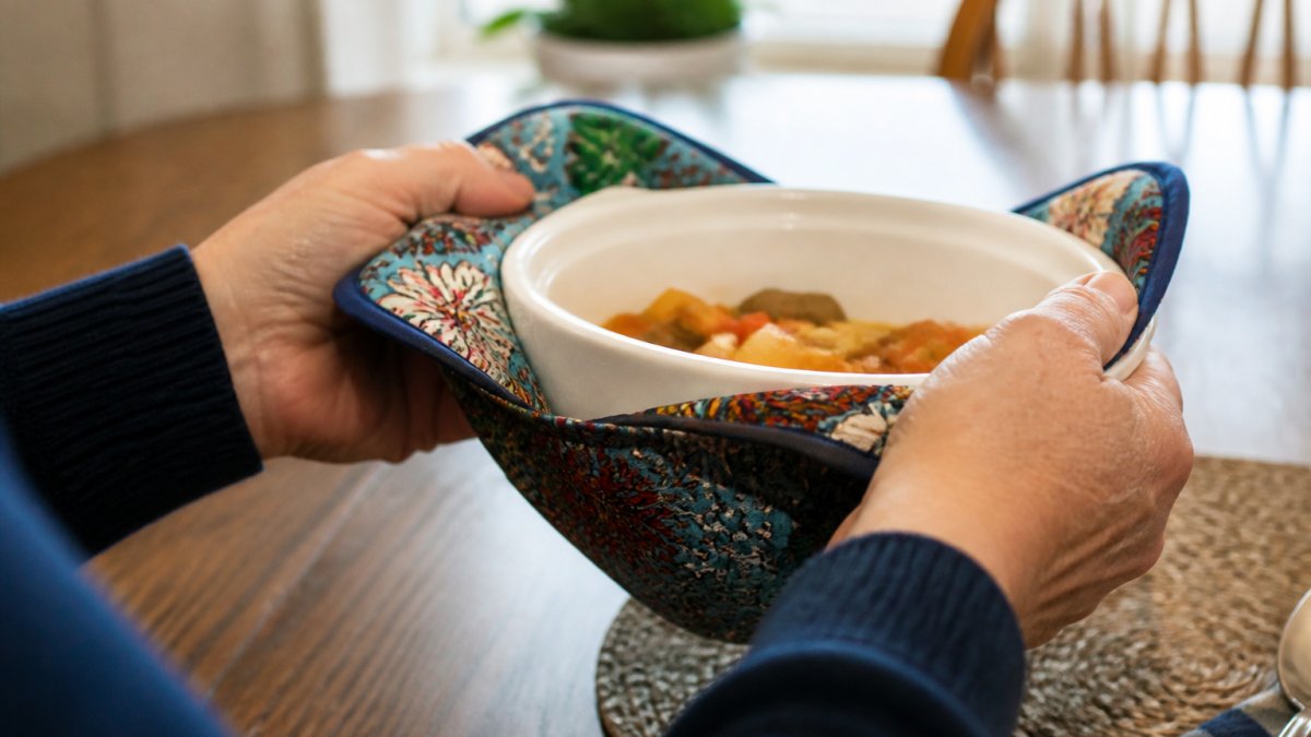 Close-up of hands holding a microwave bowl with a colorful fabric cozy, showing secure grip and heat protection during mealtime with warm lighting