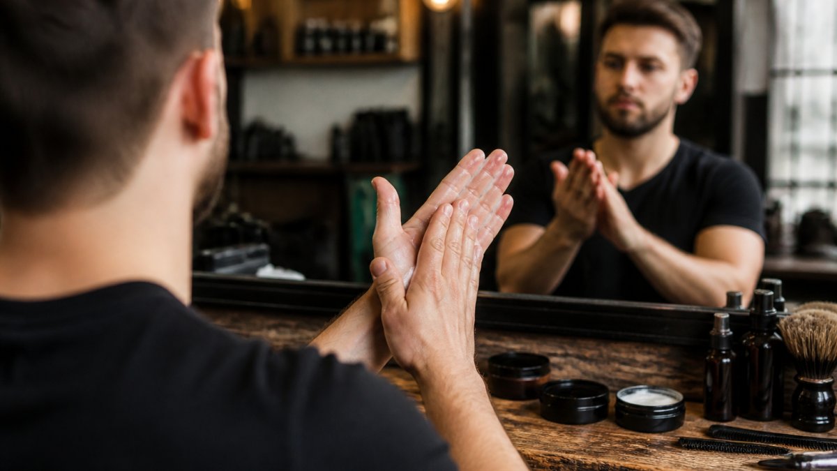 Close-up of a man's hands warming a small amount of styling paste between his palms in front of a mirror, barbershop counter with a few product jars softly blurred behind him