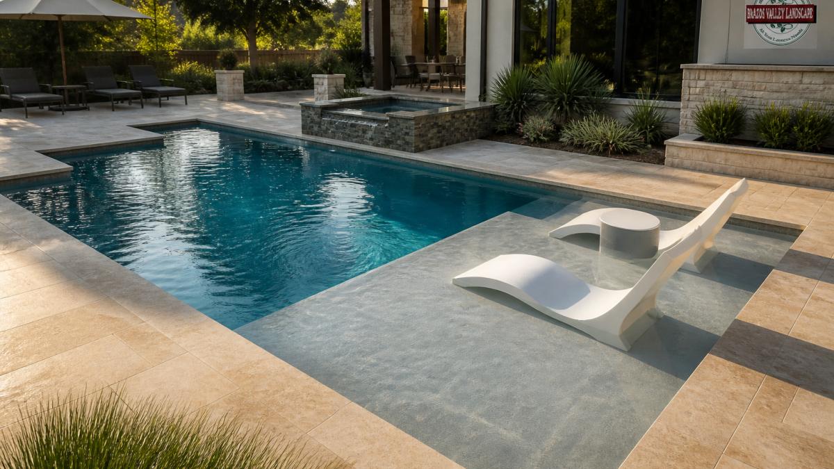 A rectangular custom swimming pool with clean geometric lines and a tanning ledge, set against a modern backyard patio with warm natural stone decking and lounge chairs under late-afternoon light