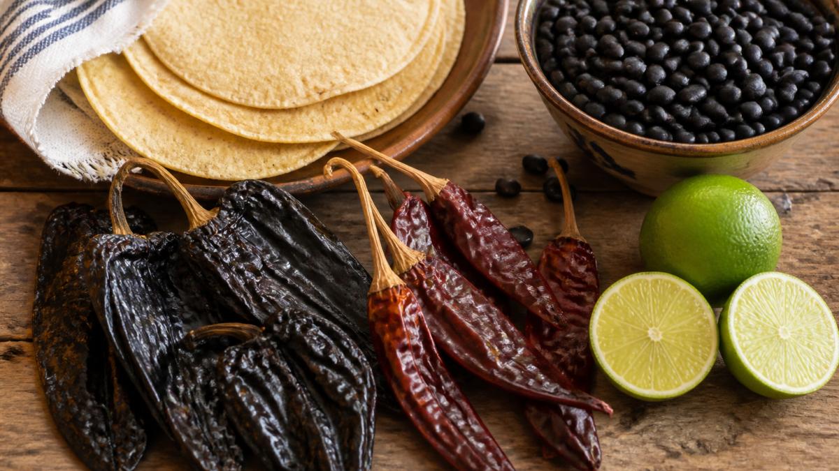 Colorful flat-lay of fresh corn tortillas, dried ancho and guajillo chiles, black beans, and halved limes arranged on a rustic wooden table with warm natural light