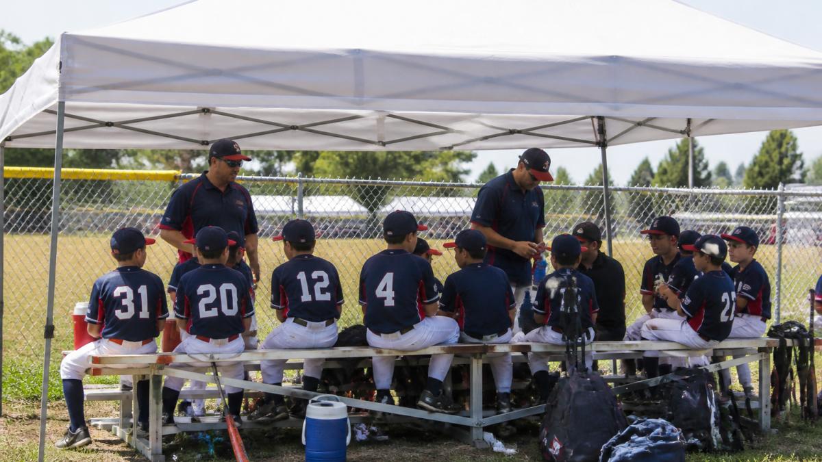 Youth baseball team gathered under a custom-branded canopy at a tournament, with players sitting on benches and coaches organizing equipment underneath the shade during a break between games