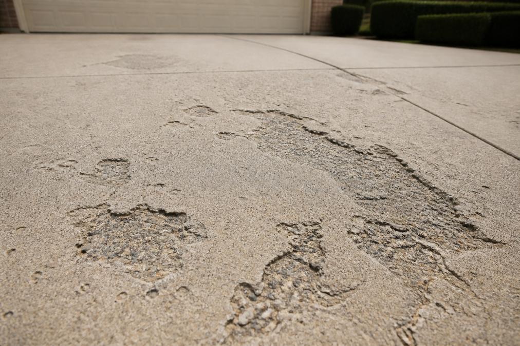 Close-up of a residential driveway surface showing visible concrete spalling, scaling, and surface flaking damage in natural daylight with varying degrees of deterioration