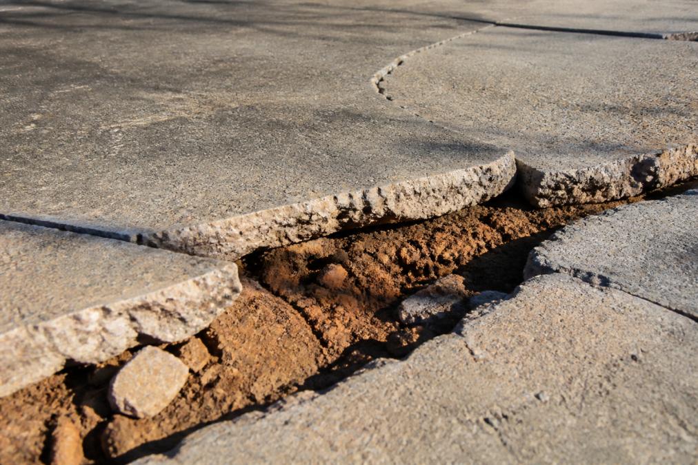 Close-up of cracked concrete driveway surface showing deep fissures and displacement, with exposed clay soil visible beneath the broken edges under bright afternoon sunlight