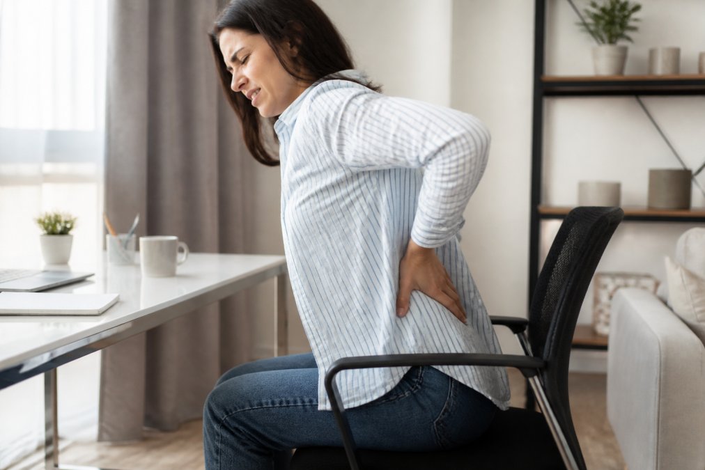 Person sitting at a desk grimacing in discomfort from sciatica pain while holding their lower back