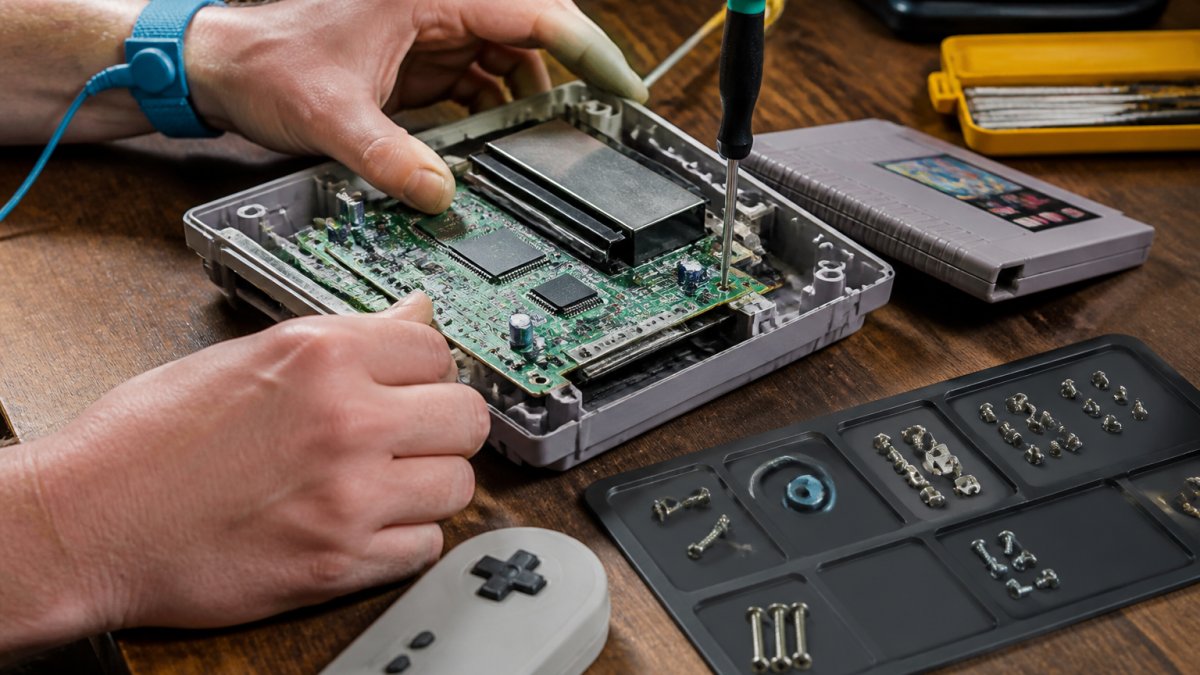 Close-up of hands in an anti-static wrist strap holding a small screwdriver over an opened retro game console on a workbench, with screws sorted on a magnetic mat and a controller and cartridge nearby