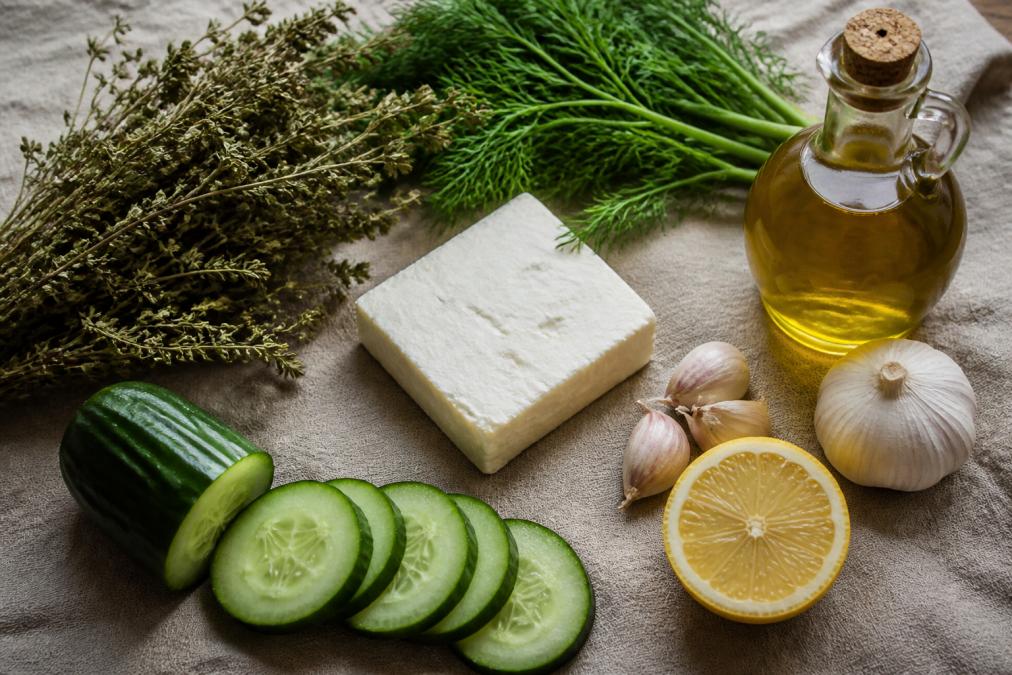 Overhead flat lay of Greek pantry staples including dried oregano bundles, fresh dill, a block of feta cheese, whole garlic cloves, a halved lemon, sliced cucumber, and a glass jar of olive oil arranged on a natural linen cloth