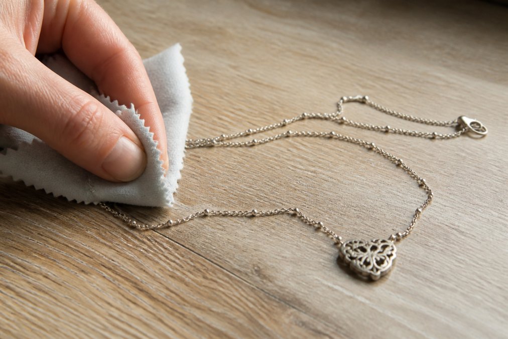 A polishing cloth gently cleaning a tarnished sterling silver necklace on a light wood surface, with soft natural light illuminating the fabric and metal
