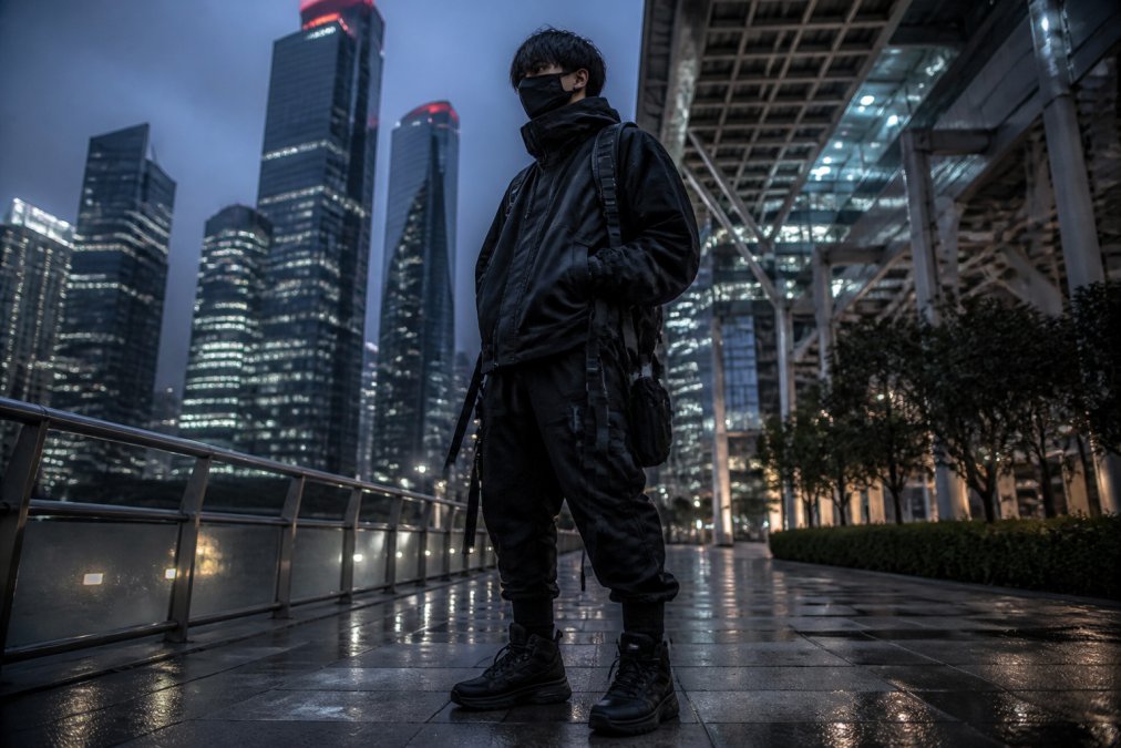 Person wearing techwear streetwear with high-collar technical jacket, cargo pants featuring multiple straps and buckles, and tactical accessories, standing in an urban megacity environment with neon signage and modern architecture at dusk