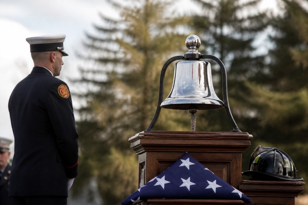 A firefighter in dress uniform standing beside a ceremonial bell during a memorial service, with a folded flag and helmet nearby, soft natural light, respectful and solemn atmosphere