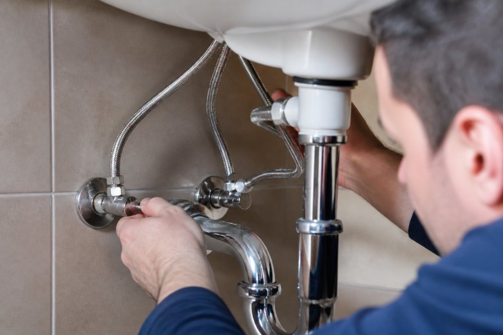 Close-up of a plumber's hands inspecting under-sink water supply lines and shutoff valves, checking for leaks after a repair in a residential bathroom