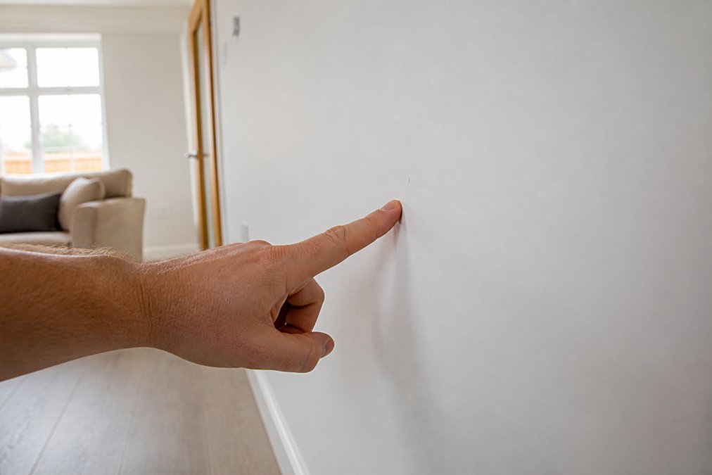 A close-up of a person's hand pointing at a small paint imperfection on a white interior wall in a bright, newly finished living room