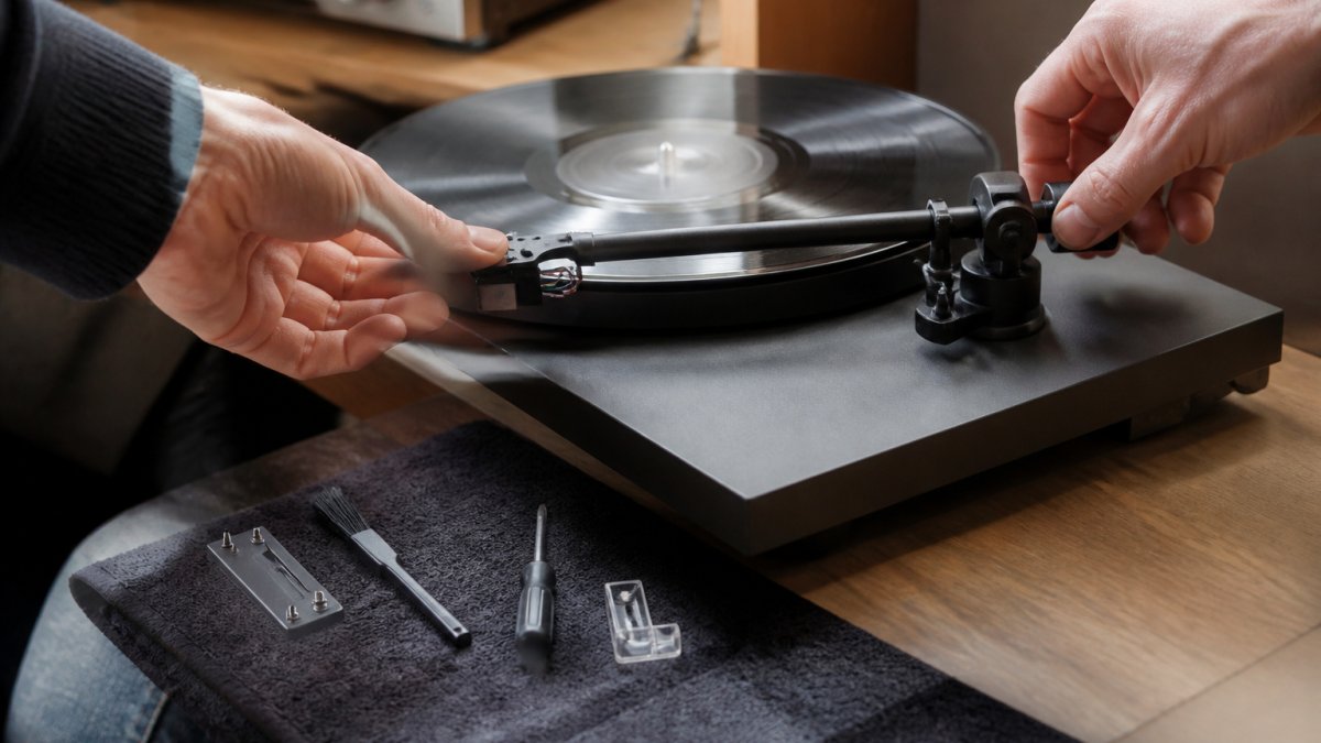 Professional hands carefully adjusting a turntable's tonearm counterweight and cartridge alignment over a vinyl record on the platter, with a stylus visible under warm studio lighting