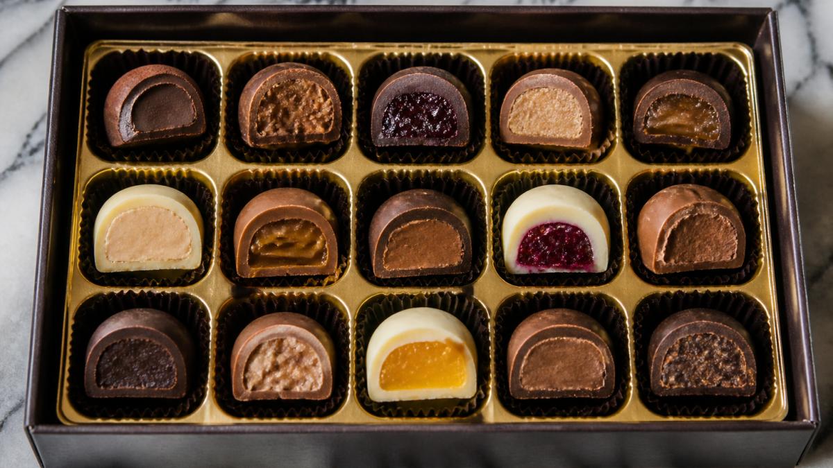 Close-up overhead shot of an open assorted chocolate box on a marble surface, showing individual chocolates cut in half to reveal ganache, praline, caramel and fruit fillings inside